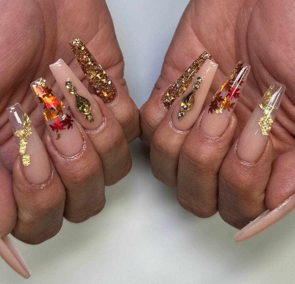 Autumn-themed nails with gold glitter, leaf designs, and jewel accents on a nude base. Perfect fall nail look created at NAB Nail Bar in Las Vegas.