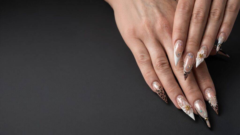 Close-up of hands with long stiletto nails featuring nude bases, white pointed tips, and delicate glittery accents with small white flowers.