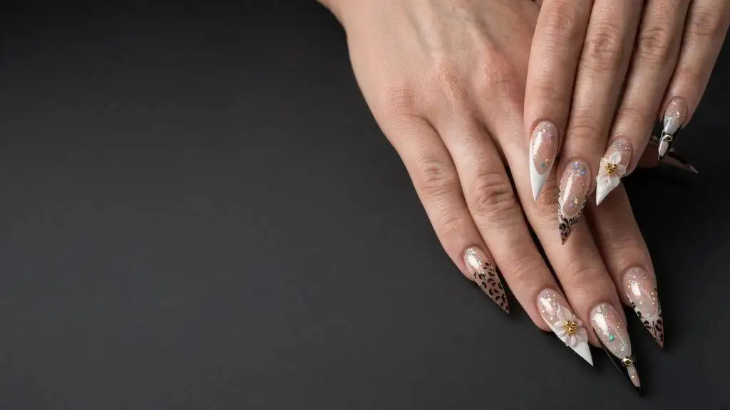 Close-up of hands with long stiletto nails featuring nude bases, white pointed tips, and delicate glittery accents with small white flowers.