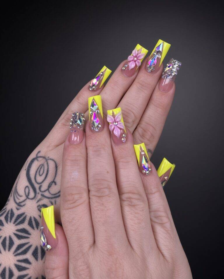 Bright yellow coffin nails with large rhinestone accents and 3D floral charm on index finger, chandelier-like shimmer on others, tattooed wrist visible behind hands