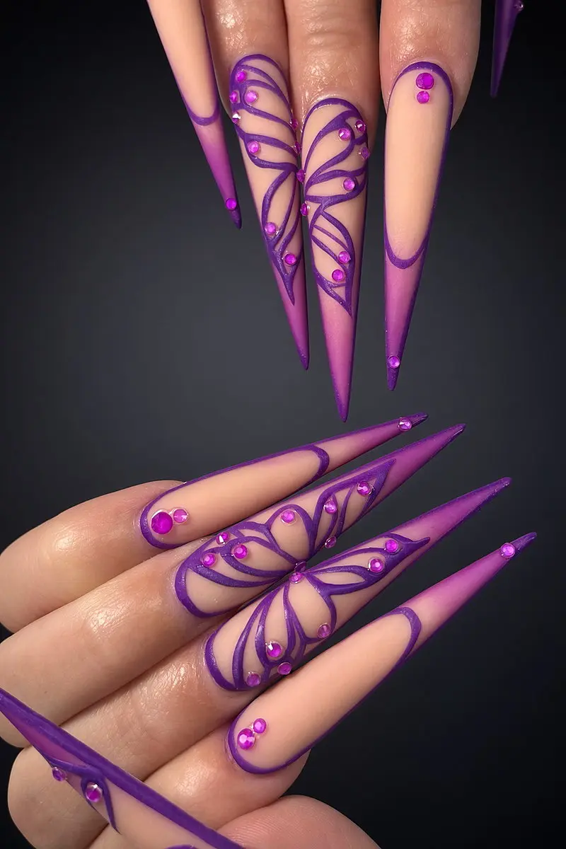Long stiletto nails with intricate purple lace like patterns and pink rhinestones on a dark background for a dramatic manicure Nab nail bar