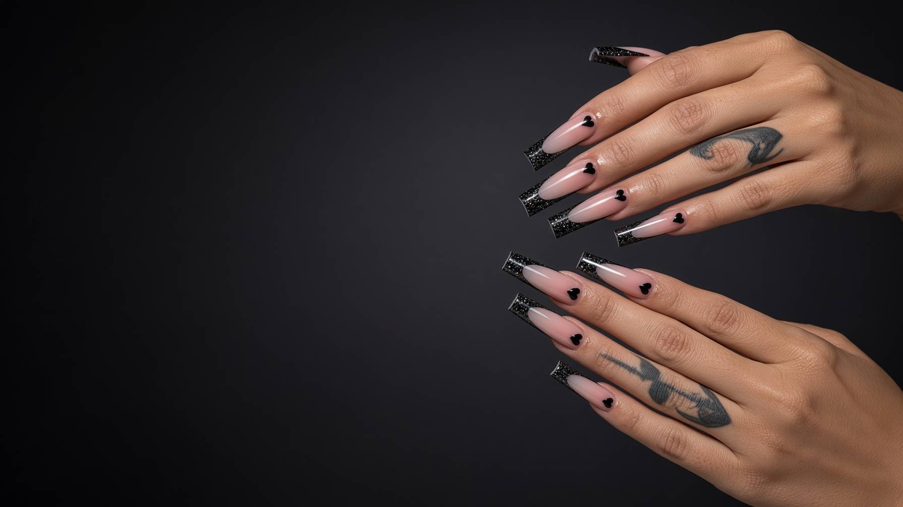 Long acrylic nails with pink to nude gradient and black heart accents at the tip glittery black french tips hands with tattoos against a dark backdrop nab nail bar | nails lashes and beauty in las vegas