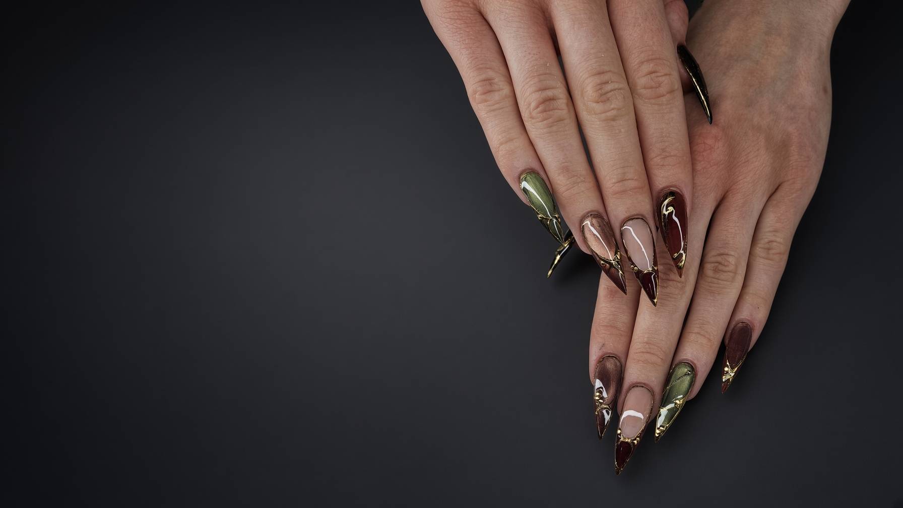 Close up of hands with long stiletto nails featuring green brown and gold geometric art against a dark background Nab nail bar | nails lashes and beauty in las vegas