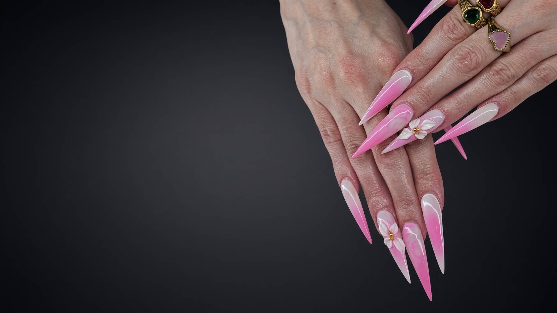 Close up of hands with long pink ombre stiletto nails flower accent and gold rings against a dark background Nab nail bar | nails lashes and beauty in las vegas