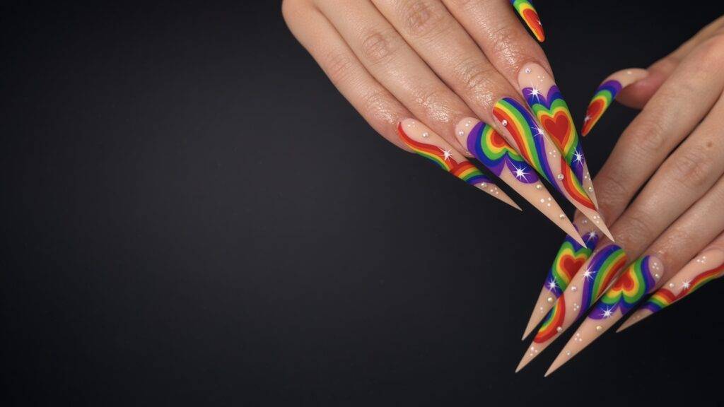 Long, pointed nails with rainbow spirals and heart shapes, adorned with small rhinestones on a dark background.
