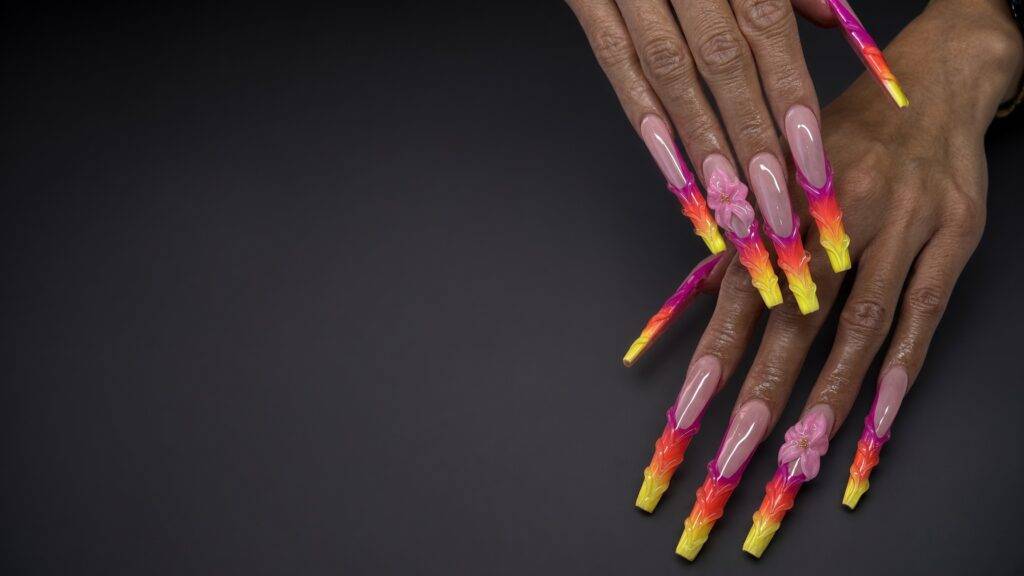 Long acrylic nails with bright pink to yellow gradient tips and embedded flower accents set against a dark backdrop nab nail bar | nails lashes and beauty in las vegas