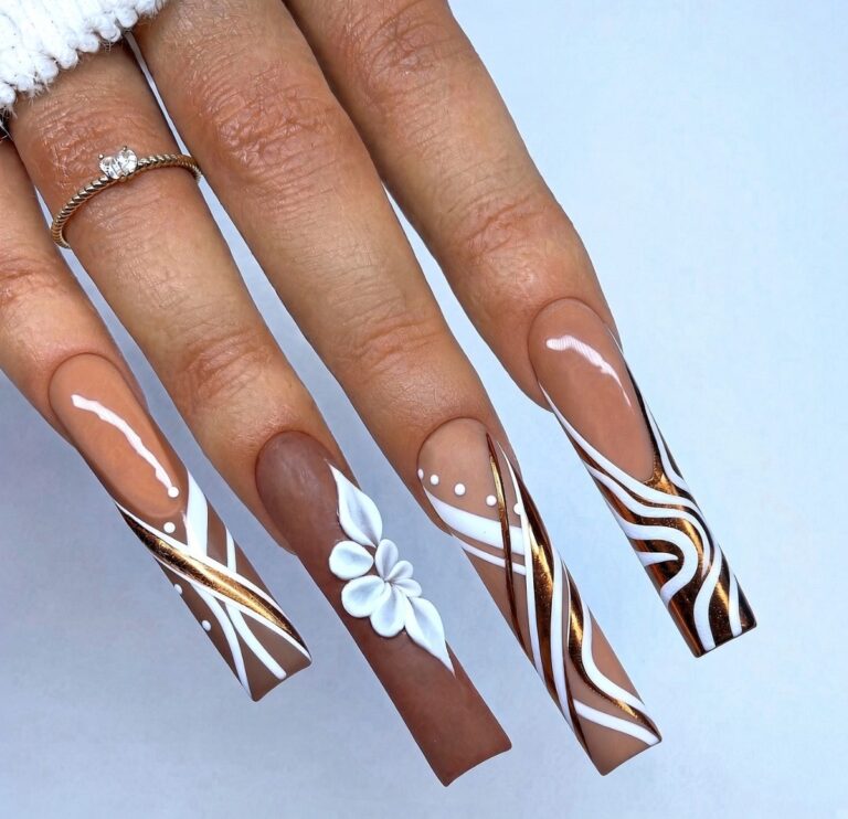Long coffin-shaped acrylic nails with brown nude base and white/gold geometric and floral nail art, hand with ring against light blue background.