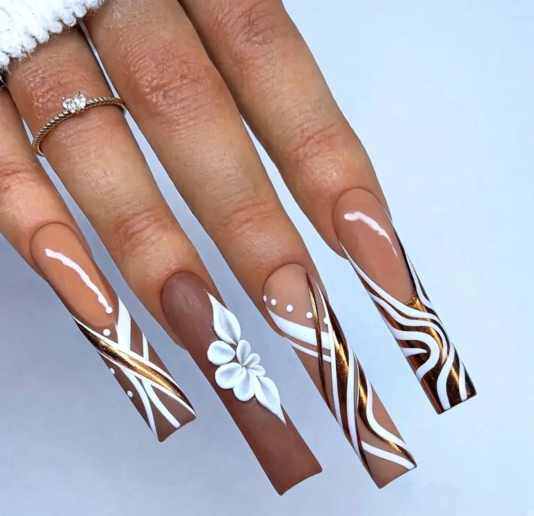 Long coffin-shaped acrylic nails with brown nude base and white/gold geometric and floral nail art, hand with ring against light blue background.