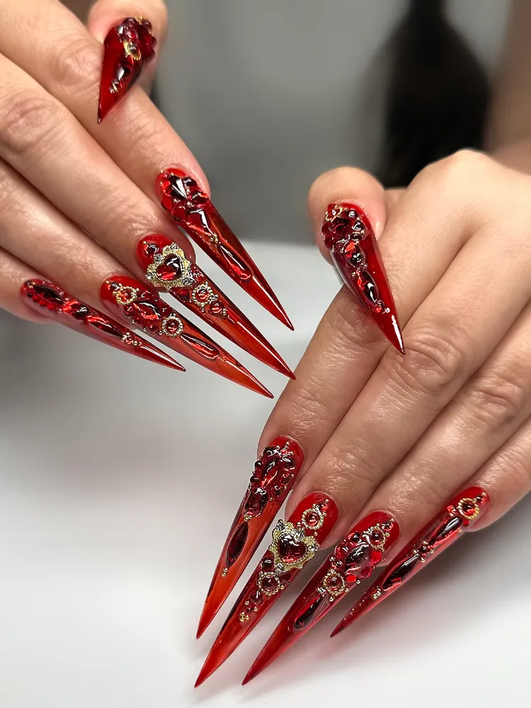 Long translucent red chrome stiletto nails with gold heart charms and red gems for a holiday party at NAB Nail Bar Las Vegas