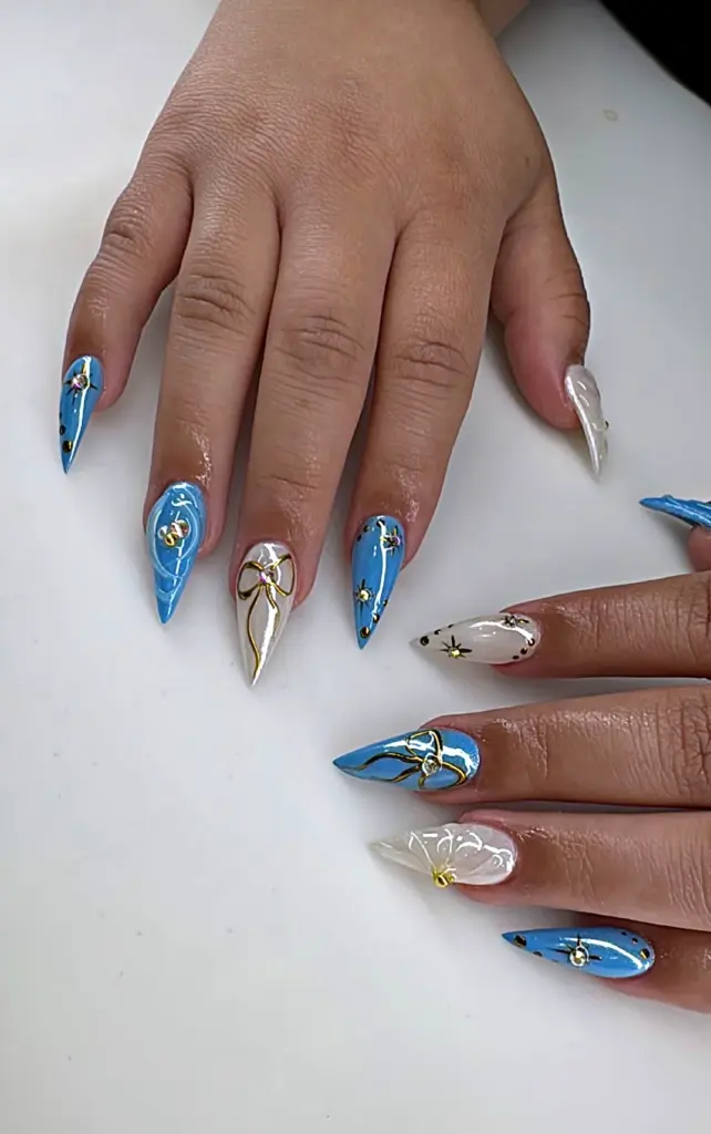 Short stiletto Gel X nails in blue and pearl white with gold bows and crystals at NAB Nail Bar in Las Vegas